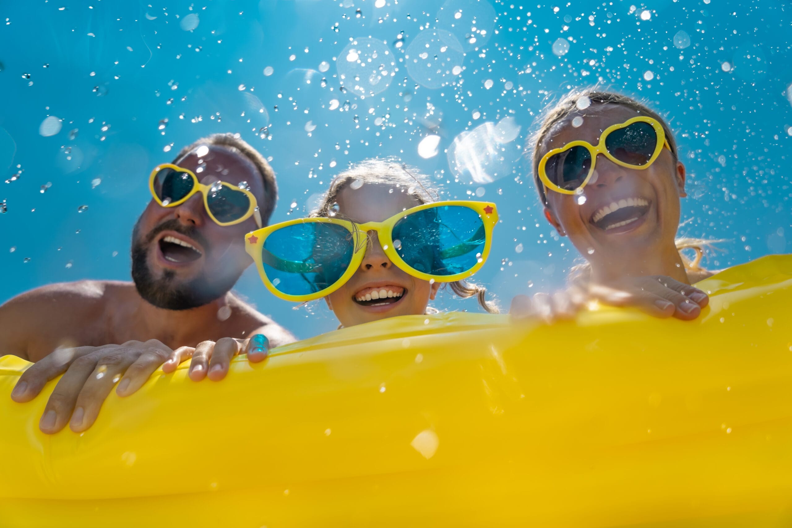 Man, young girl and women laughing with big yellow glasses with water splashing around them on a yellow floating device