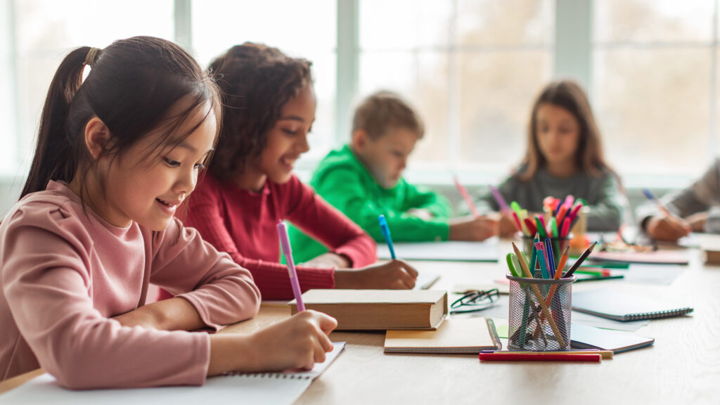 Schoolgirl Taking Notes Writing Essay Learning In Modern Classroom At School. Multiracial Children Having Class. Knowledge And Education. Selective Focus, Panorama